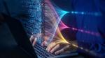 Een zilverkleurige laptop met zwarte toetsen, handen op een toetsenbord, met daaroverheen een keur aan kleurrijke digitale code die naar technologie verwijst.