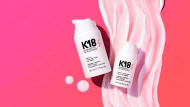 Twee witte flessen K18-haarbehandeling op een roze achtergrond met golvende crème.