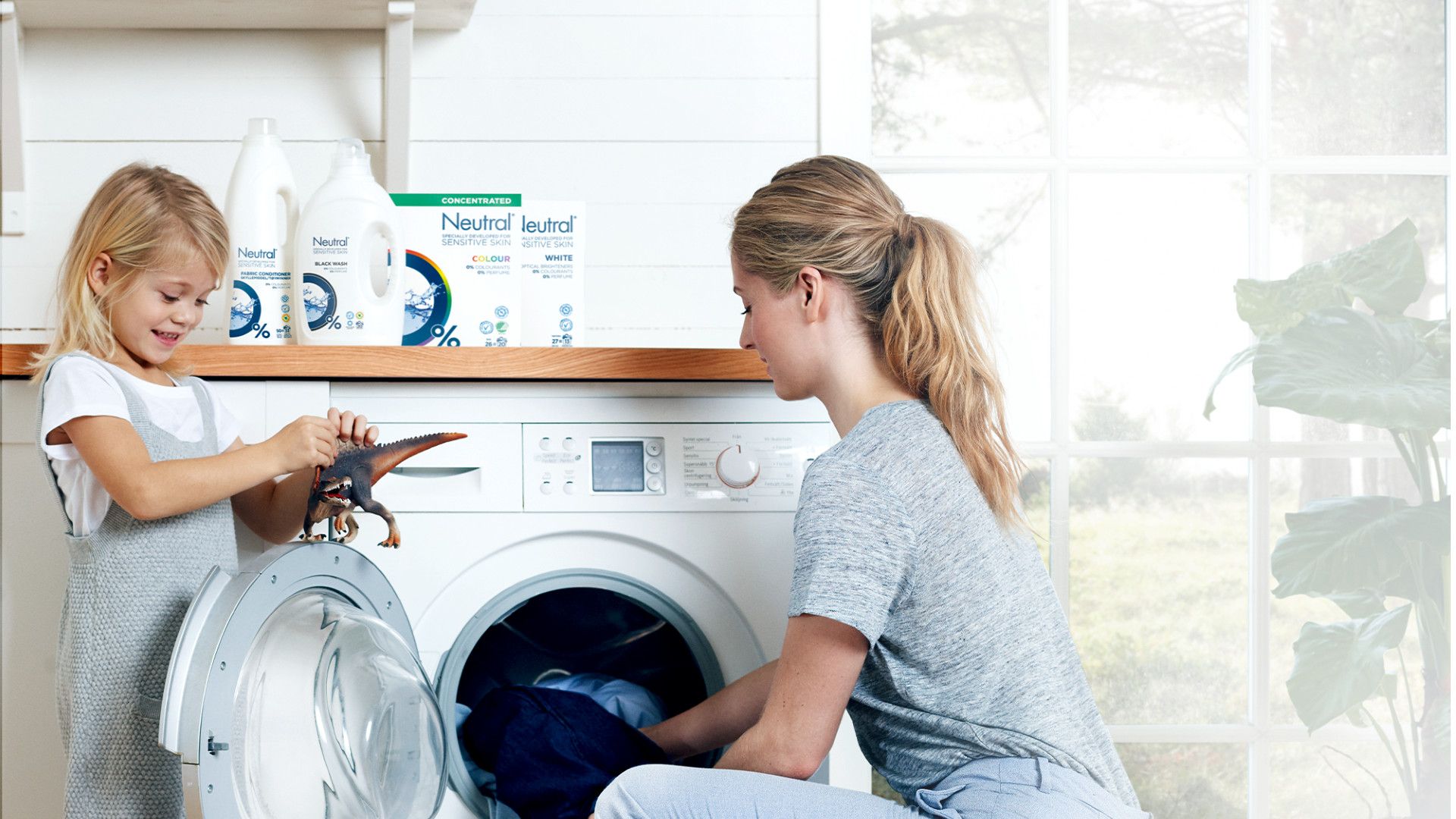 Een vrouw stopt de was in de wasmachine, terwijl een kind in de buurt een speelgoeddinosaurus vasthoudt. Op de achtergrond staan ​​wasmiddelen op een plank.