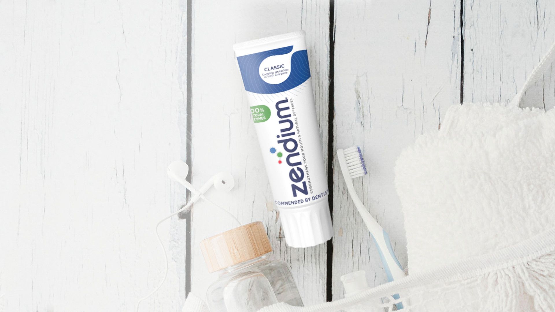 Een afbeelding van mondhygiëneartikelen, waaronder Zendium-tandpasta, een tandenborstel, een glazen potje en witte oordopjes, plat op een houten ondergrond.