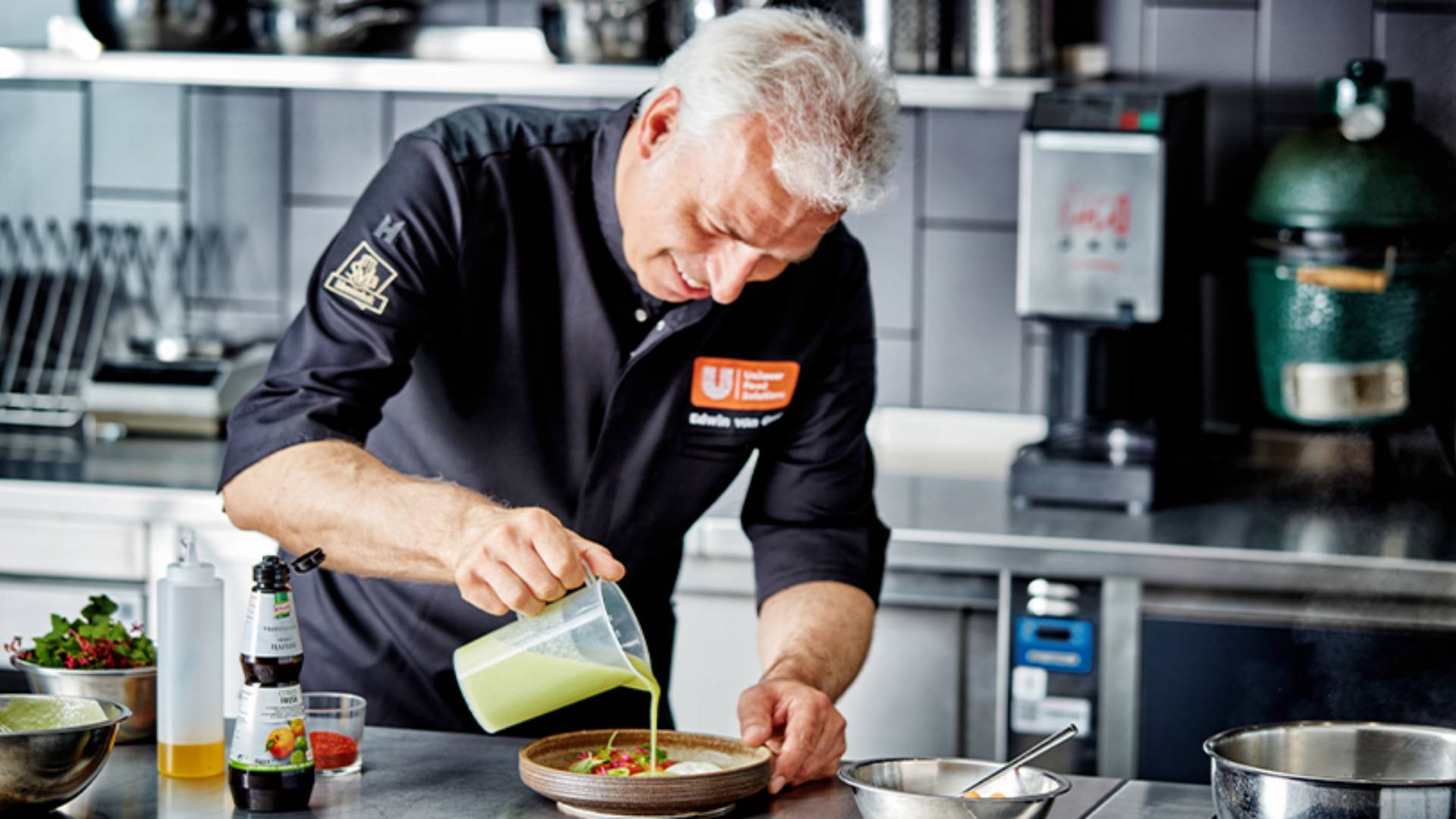 Een mannelijke chef staat in de keuken en giet saus over een gerecht.