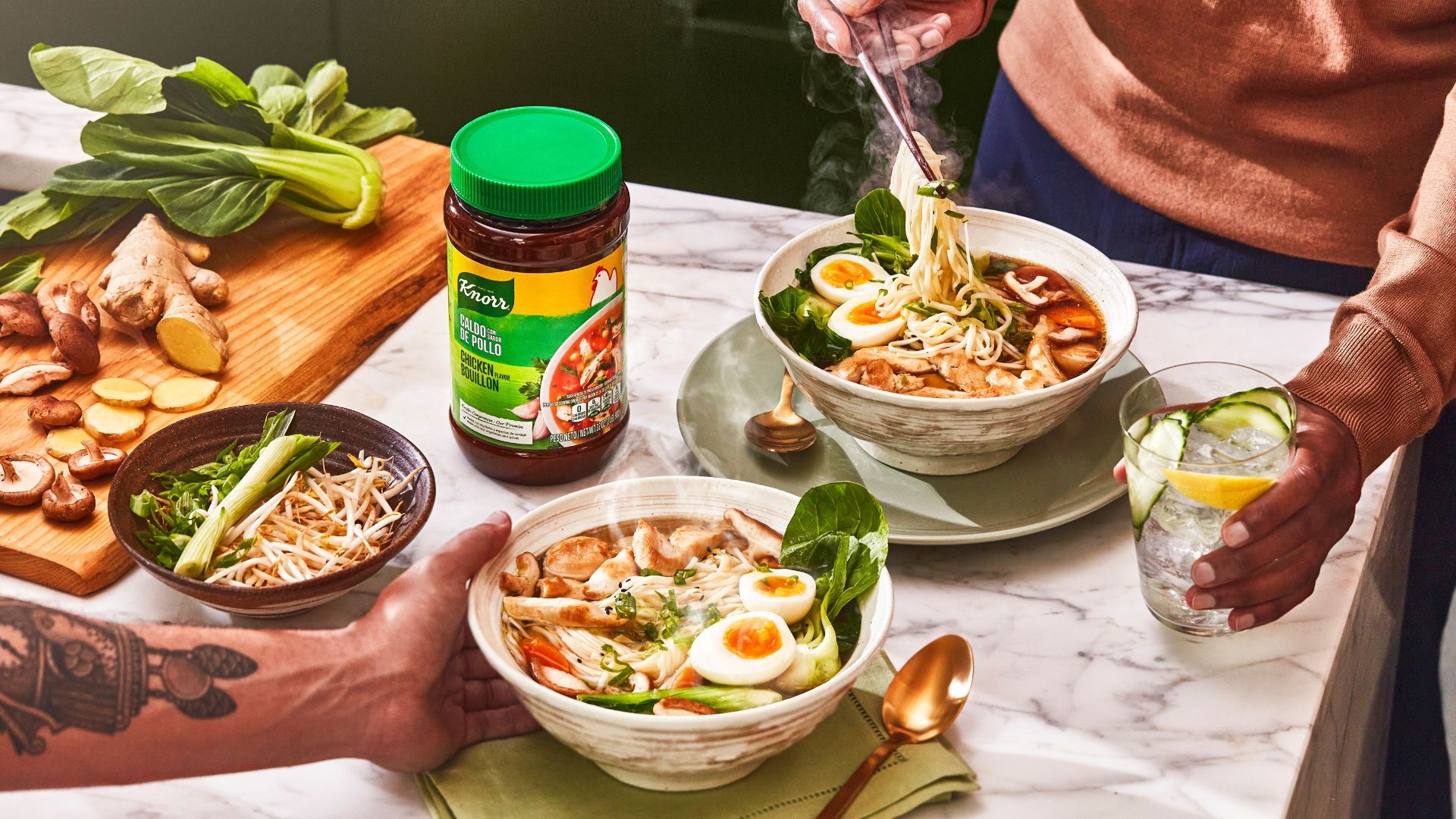 Een tafel gedekt met twee kommen met ramennoedels, een snijplank met ingrediënten en een pot met Knorr-kippenbouillon.